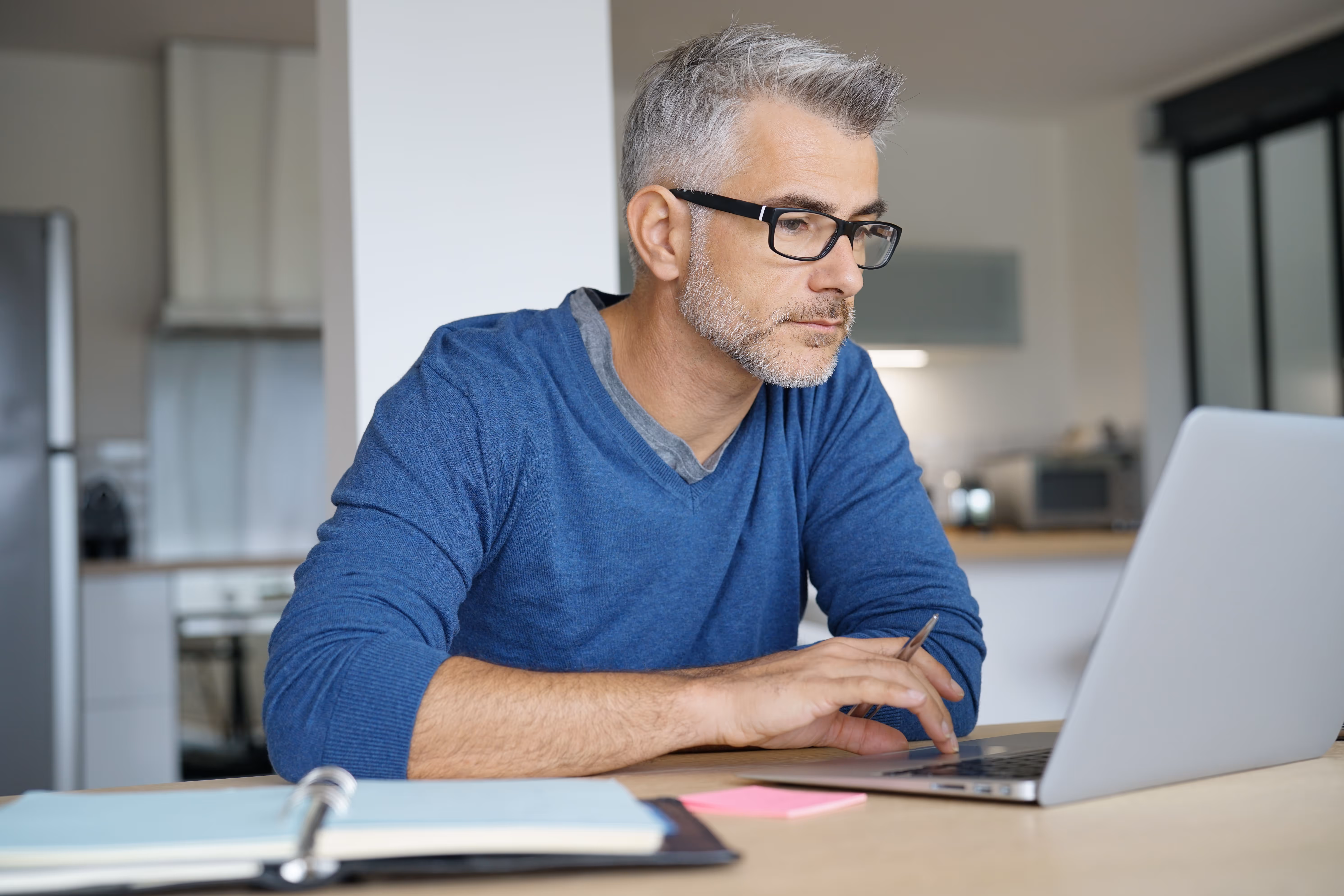 Middle-aged man with gray hair and glasses working on a laptop at a kitchen table, with a binder and sticky notes in front of him.